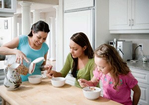 Three females sitting in the kitchen eating cereal out of white bowls.